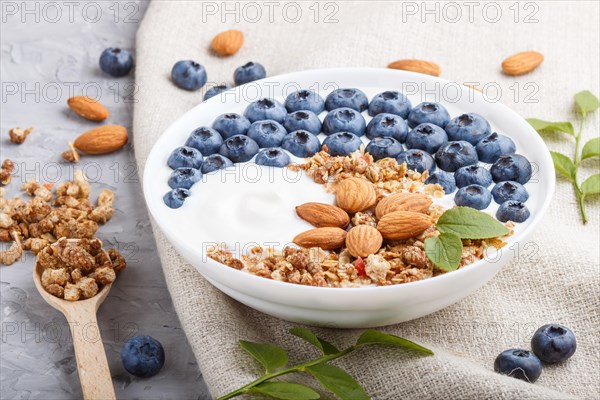 Yoghurt with blueberry, granola and almond in white plate with wooden spoon on gray concrete background and linen textile. side view, close up