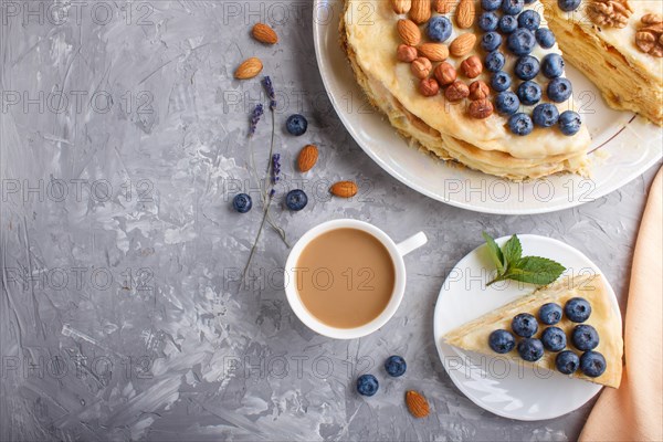 Homemade layered Napoleon cake with milk cream. Decorated with blueberry, almonds, walnuts, hazelnuts, mint on a gray concrete background and cup of coffee. top view. flat lay, copy space