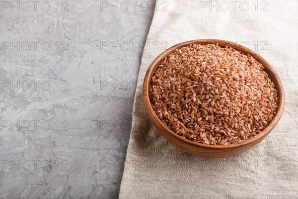 Wooden bowl with unpolished brown rice on a gray concrete background and linen textile. Side view, copy space