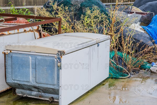 Old abandoned refrigerator laying on wet concrete in front of weeds and other debris on rainy morning in South Korea