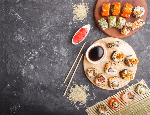 Different kinds of maki sushi rolls with salmon, sesame, cheese, roe and chopsticks, soy sauce, marinated ginger on a black concrete background. Top view, flat lay, copy space. Japanese food concept