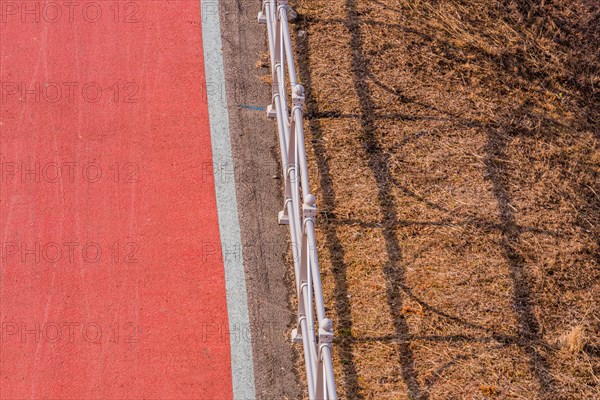 Looking down onto red bike path next to metal guardrail casting its shadow on brown grassy area in South Korea