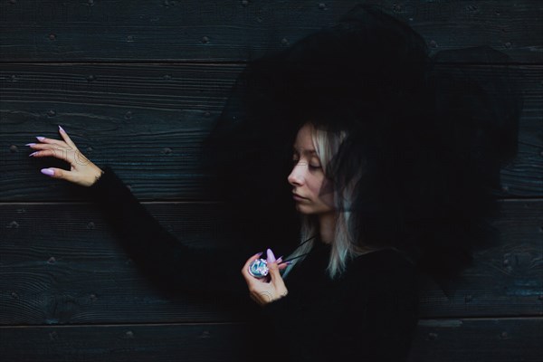 Female white blonde model wear black furry hat leaning by her side against a wooden wall, holding an object thoughtfully