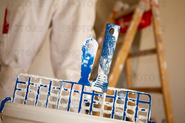 Symbolic image of painting work: Close-up of a paint bucket with scraper grid, in the background a painter's ladder