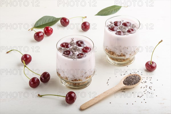 Yoghurt with cherries, chia seeds and granola in glass with wooden spoon on white wooden background. side view, close up