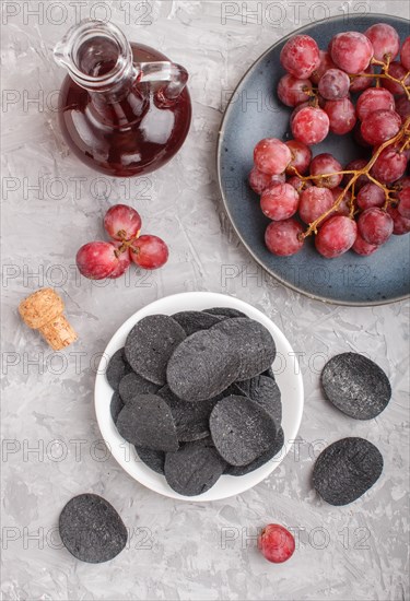 Black potato chips with charcoal, balsamic vinegar in glass, red grapes on a blue ceramic plate on a gray concrete background. Top view, flat lay, close up