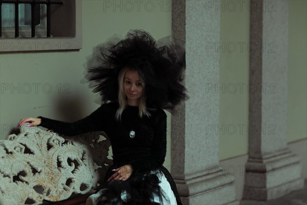 A woman on a bench by a historical building, her ornate hat adding to the scene's melancholy