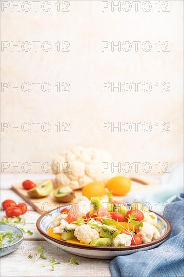 Vegetarian salad of cauliflower cabbage, kiwi, tomatoes, microgreen sprouts on white wooden background and blue linen textile. Side view, copy space, selective focus