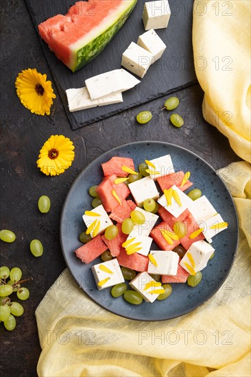 Vegetarian salad with watermelon, feta cheese, and grapes on blue ceramic plate on black concrete background and yellow linen textile. top view, close up, flat lay