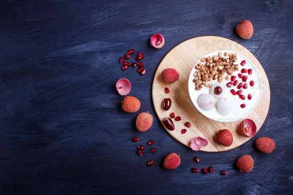 Greek yogurt with lychee, pomegranate seeds and granola in a white plate on a black wooden background, top view, flat lay, copy space