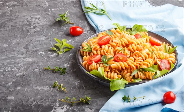 Fusilli pasta with tomato sauce, cherry tomatoes, lettuce and herbs on a black concrete background with blue textile. side view, copy space