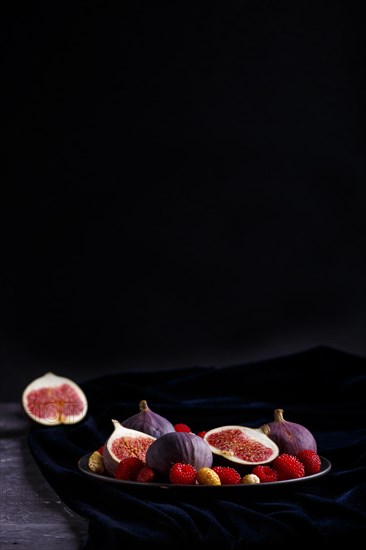Fresh figs, strawberries and raspberries on blue ceramic plate on black concrete background and blue velvet textile. side view, copy space, selective focus, low key
