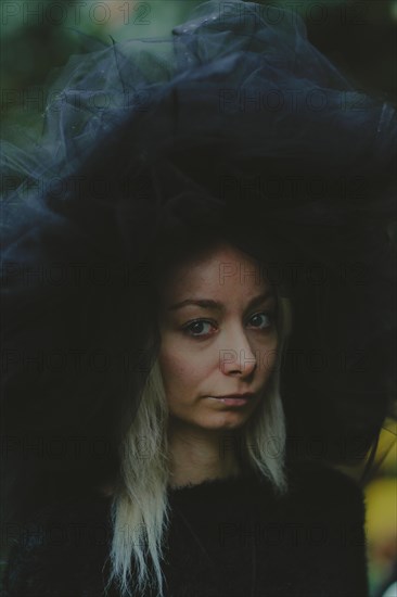 A woman wearing a tulle hat appears thoughtful, captured outdoors in natural lighting