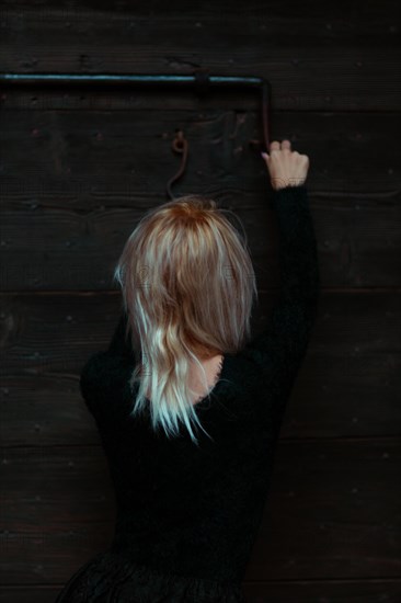 Back view of a blonde thin white woman dressed in black reaching up to a rusty antique metal lock on a wooden door