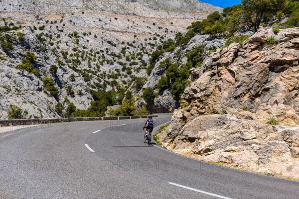 A cyclist rides along a winding mountain road surrounded by imposing rock faces under a clear blue sky, Majorca