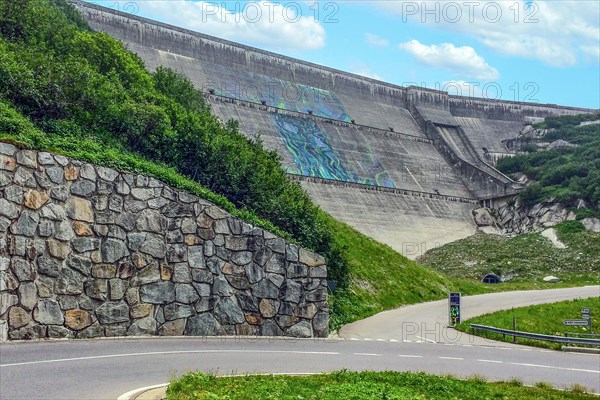 View from road pass road alpine road to road level below in front of outside of large dam wall of reservoir Lac du Mont Cenis Mont-Cenis reservoir next to alpine pass mountain pass Col du Mont Cenis in French High Alps, Departement Savoie, Region Auvergne-Rhone-Alpes, France, Europe