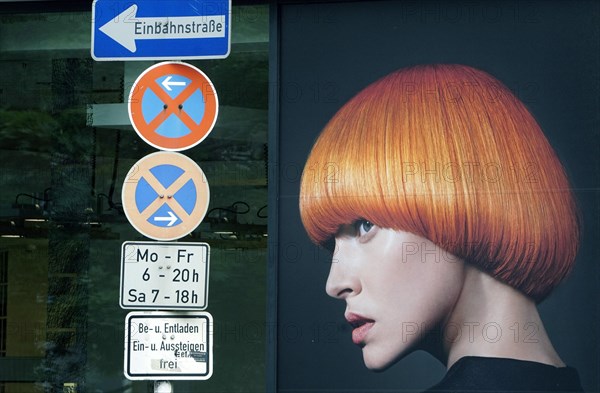 An advertising poster in the window of a hairdressing salon shows a photo of a woman with red hair. It looks as if the woman is looking at a forest of traffic signs, 17.06.2019