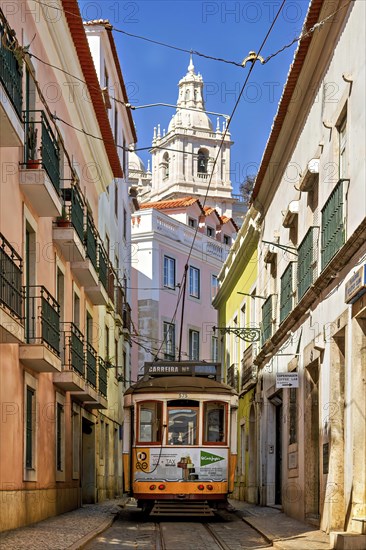 The historic yellow tram winds its way through the historic centre of Lisbon past the old houses of this beautiful city. In the background is a typical Portuguese church