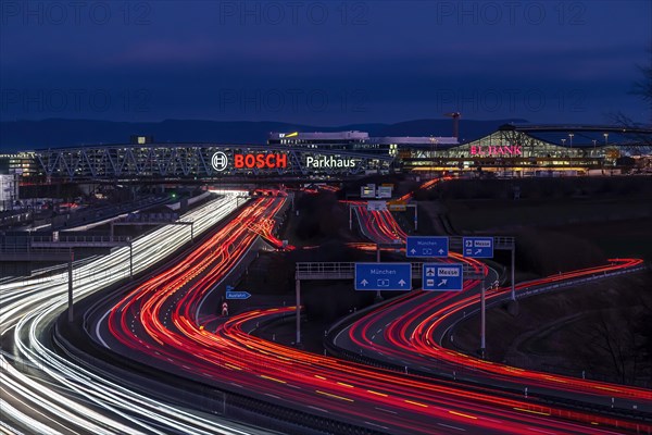 A8 motorway at Stuttgart Airport with Bosch multi-storey car park in the evening, cars create light trails. The 440 metre long construction offers space for 4200 vehicles. Bosch owns the naming rights, the stylised ignition anchor measures 12 metres in diameter. This makes it one of the largest neon signs in the world. Stuttgart, Baden-Wuerttemberg, Germany, Europe