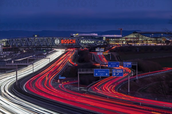 A8 motorway at Stuttgart Airport with Bosch multi-storey car park in the evening, cars create light trails. The 440 metre long construction offers space for 4200 vehicles. Bosch owns the naming rights, the stylised ignition anchor measures 12 metres in diameter. This makes it one of the largest neon signs in the world. Stuttgart, Baden-Wuerttemberg, Germany, Europe