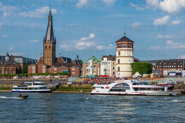 A busy stretch of river with boats and a crowd of people against the backdrop of a town with a church, Rhine embankment promenade, Duesseldorf, North Rhine-Westphalia