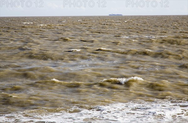 Brown choppy sea carrying sediment in suspension with distant container ship, North Sea, Suffolk, England, United Kingdom, Europe