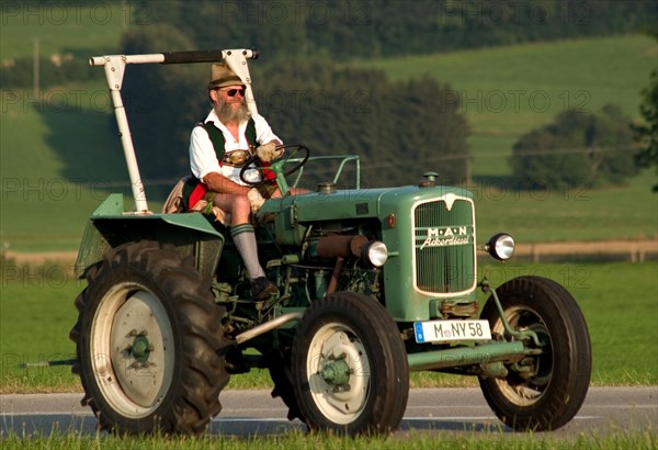 Traditionally dressed farmer in lederhosen and Tyrolean hat on a tractor, Dietramszell, Bavaria, Germany, vintage, retro, Europe