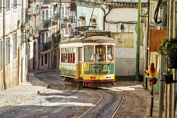 The historic yellow tram winds its way through the historic centre of Lisbon past the old houses of this beautiful city. A young woman in a yellow shirt enriches the scene