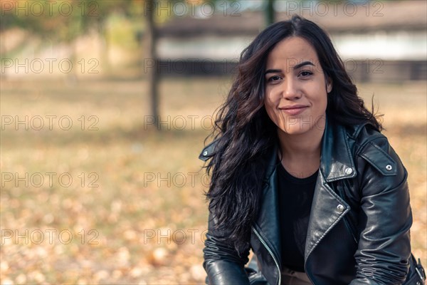 Content latina woman in a leather jacket with a relaxed posture in an autumnal park setting, outdoors, blurred background with bokeh, daytime, AI generated