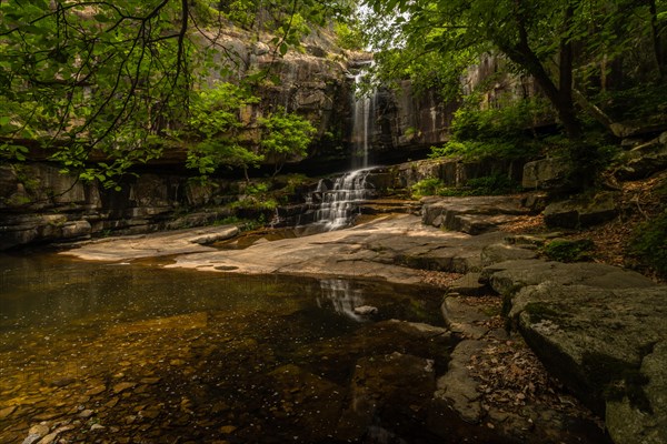 A secluded waterfall spills over ancient rocks into a still pool, shaded by a wooded canopy, in South Korea