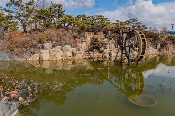 A water wheel beside a peaceful pond with rock formations and clear reflections, in South Korea