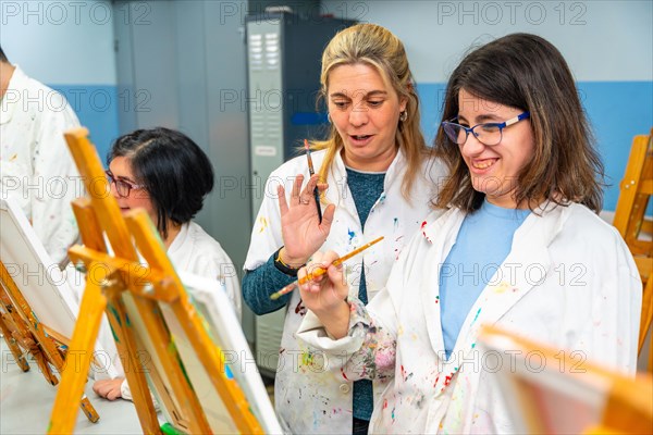 Happy woman with special needs painting in a canvas next to a teacher in the art class
