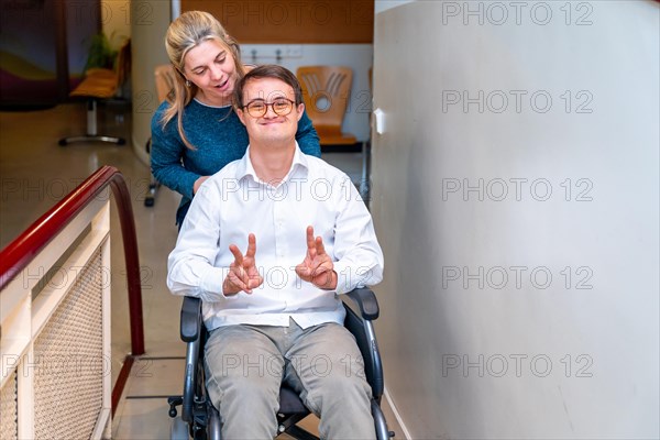 Health worker pushing a happy man with down syndrome using wheelchair while he gesturing success with fingers