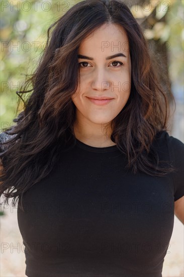 Content Cheerful hispanic young woman in a black shirt outdoors with relaxed posture and a serene look in natural light, selective focus, blurred background with bokeh, daytime, AI generated