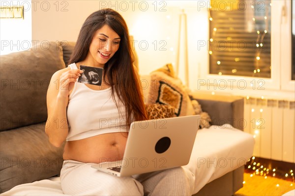 Happy pregnant woman showing ultrasound during online video call sitting on the sofa at home at night
