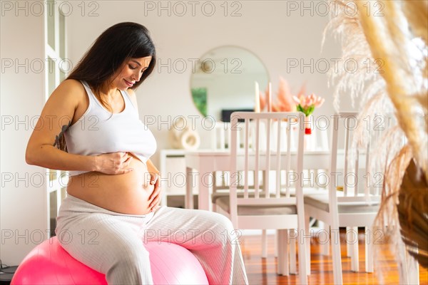 Beauty pregnant woman in sportive pregnancy clothes sitting on pilates ball at home