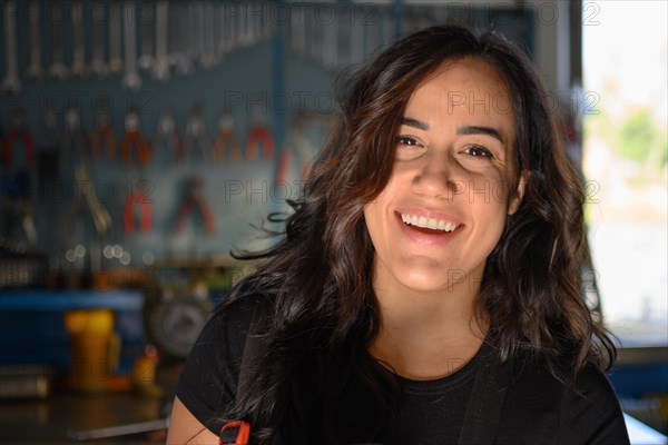 Smiling hispanic female mechanic in workshop standing in front of a workbench, a complete tool panel in background with bokeh effect, traditional male jobs by Mixed-race latino woman