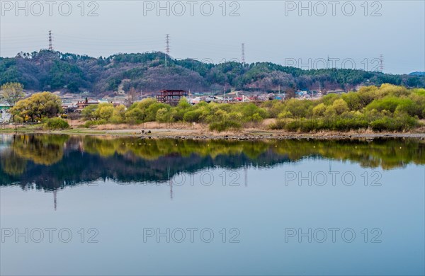 Greenery and distant buildings reflected on water with hills and cloudy skies above, in South Korea
