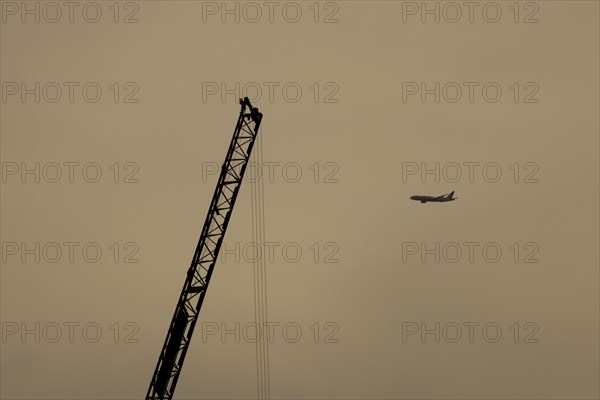 Aircraft in flight with an industrial crane in the foreground at sunset, London, England, United Kingdom, Europe