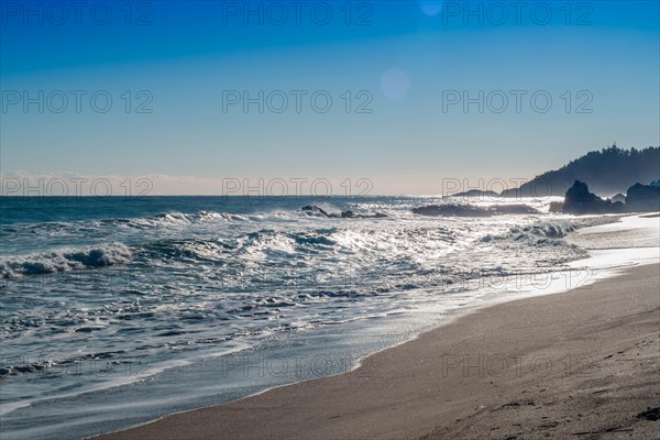 Sunlit waves crash against a rocky shore with glistening sea foam, in South Korea