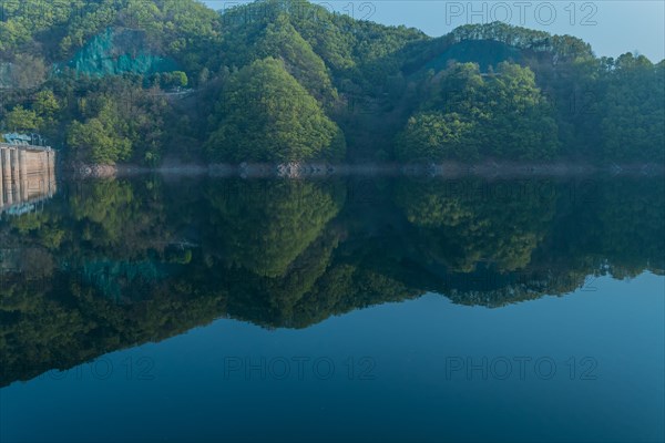 Calm reservoir with forested hills and morning mist, creating a mirror-like reflection, in South Korea