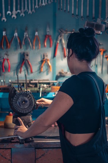 Female hispanic Mechanic working at a bench in a well-equipped workshop from behind, a complete tool panel in background with bokeh effect, traditional male jobs by Mixed-race latino woman