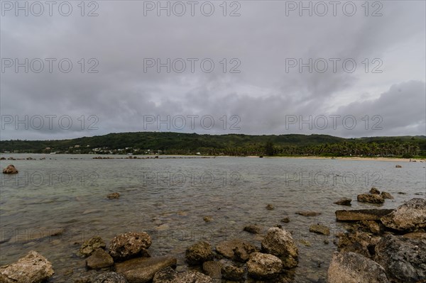 Seascape of rocky shoreline on a cloudy day with buildings on tree lined shore in the distance in Guam