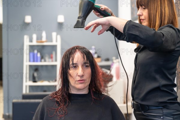 Frontal photo of a serious and patient woman sitting on hair salon while hairdresser drying her hair