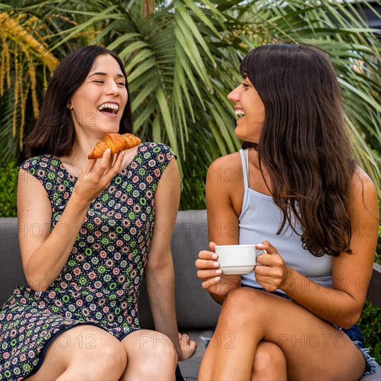 Two women are sitting on a couch, one of them is holding a croissant and the other is holding a cup of coffee. They are both smiling and laughing, enjoying each other's company
