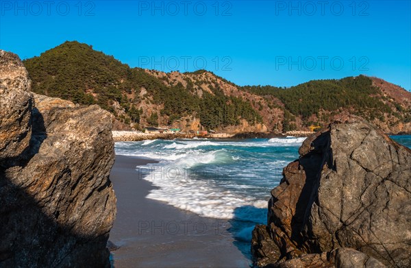 The ocean's waves crash against a cliffside with forested hills under the clear sky, in South Korea