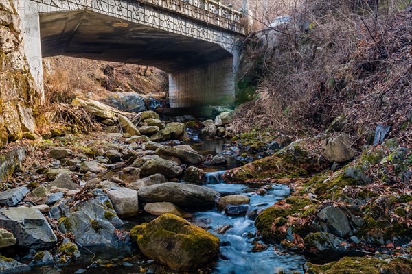 Leafless trees and rocks surround a stream flowing under a bridge in an autumn setting, in South Korea