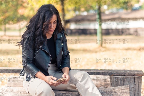 Cheerful hispanic young Woman Concentrated woman seated outdoors in fall season, texting on a smartphone, in autumn, blurred background with bokeh, daytime, AI generated