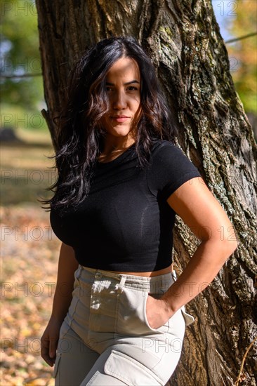 Confident hispanic sexy woman in black top and khaki pants standing by a tree in autumn, blurred background with bokeh, daytime, AI generated