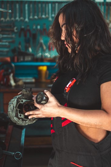 Focused Young confident skilled hispanic sexy female mechanic inspecting a car alternator in an auto repair shop, a complete tool panel in background with bokeh effect, traditional male jobs by Mixed-race latino woman